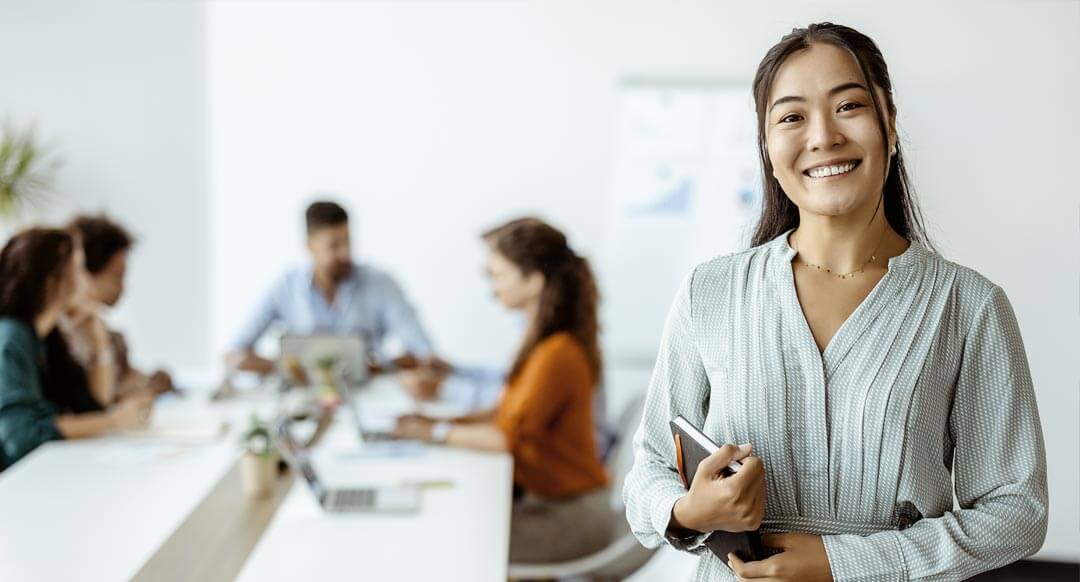 Confident young Asian businesswoman standing smiling at the camera in a boardroom with colleagues in the background